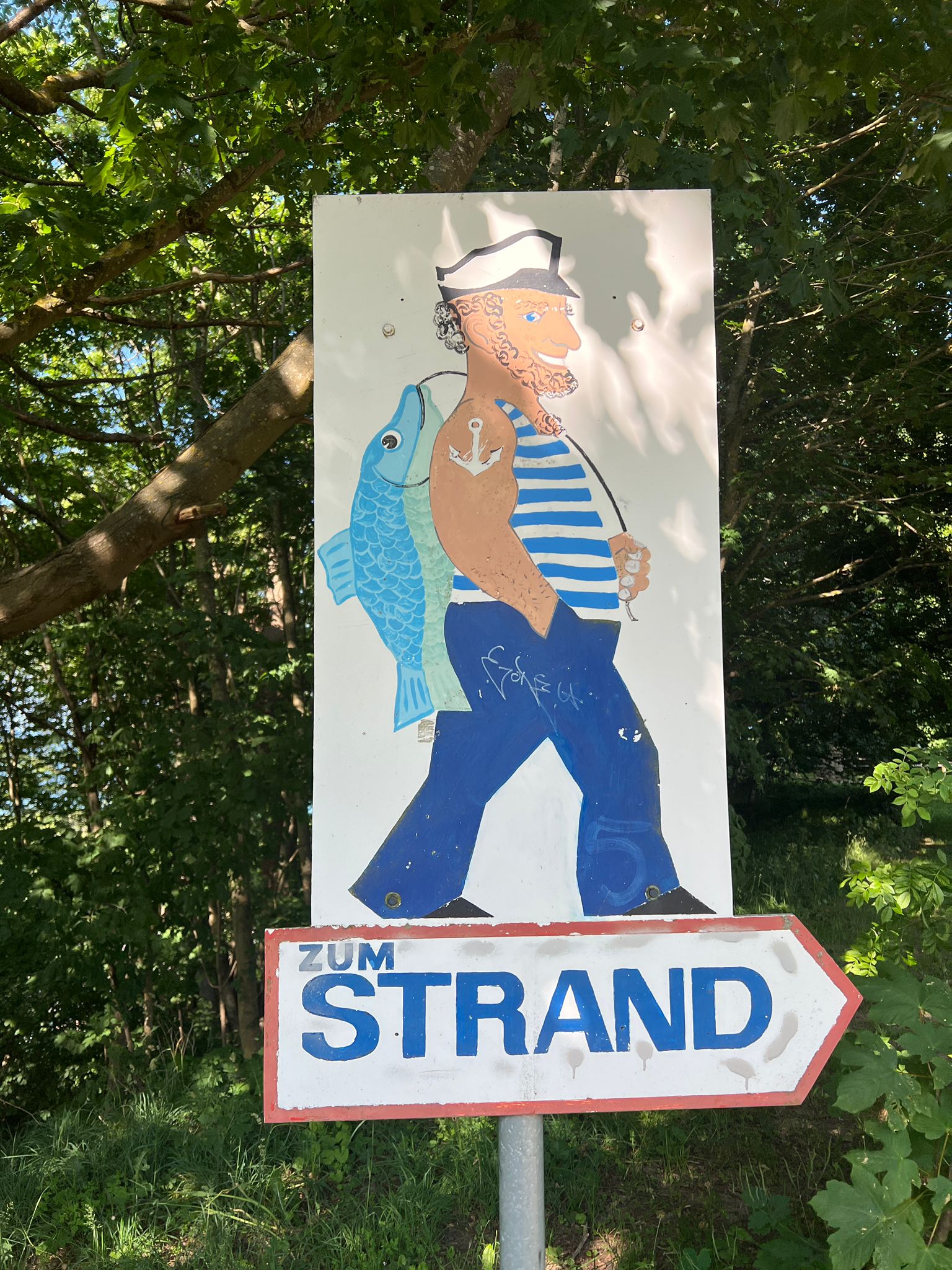 Schildverweis in der Waldpromenade zum Strand mit einem Fischer